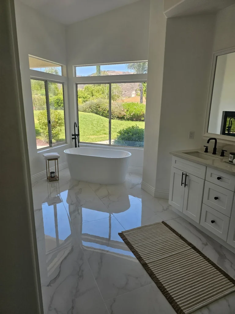 Freestanding white bathtub beside large windows, with a white vanity and veined floor tile.