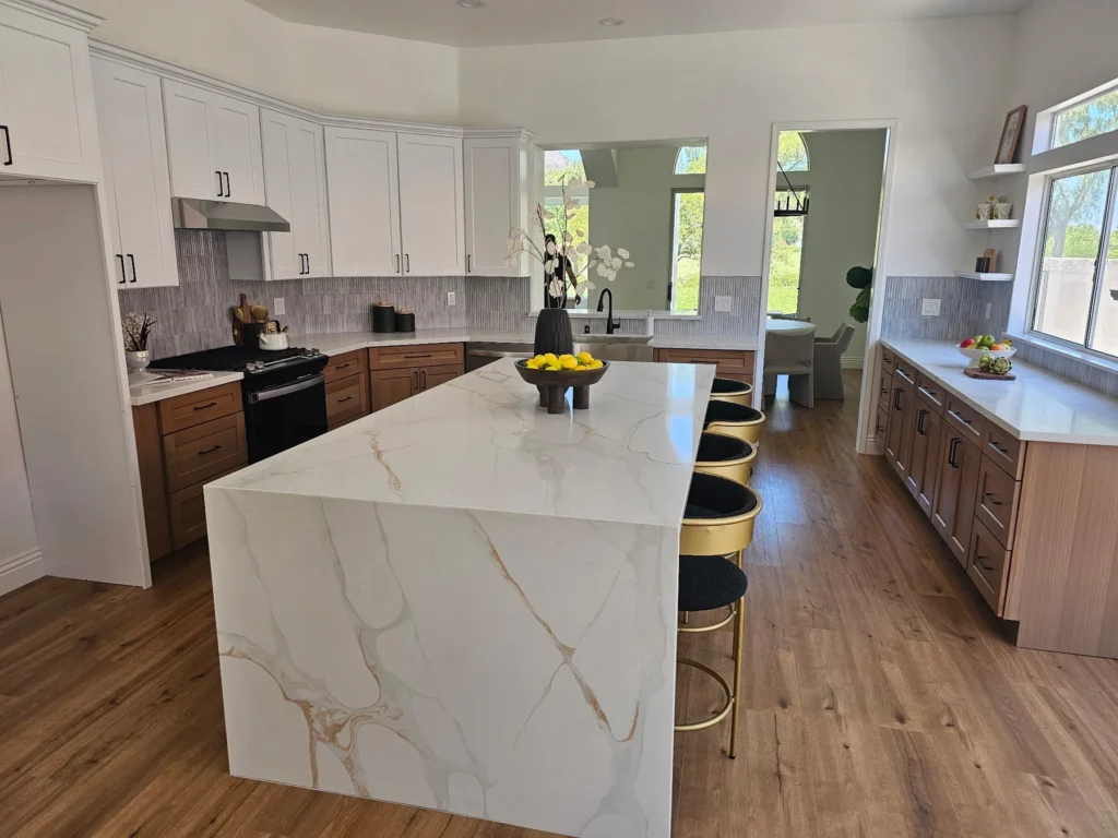Veined white waterfall countertop with seating beside wood cabinets and large kitchen windows.